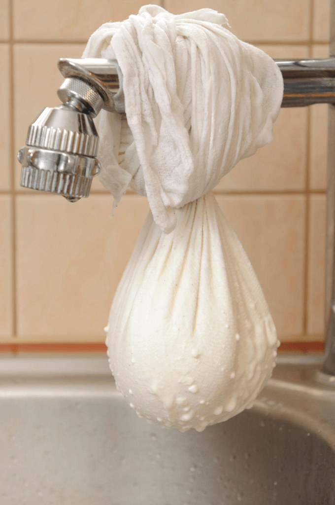 A muslin cloth is tied into a bundle and hung over the kitchen sink for straining curd to make hung curd